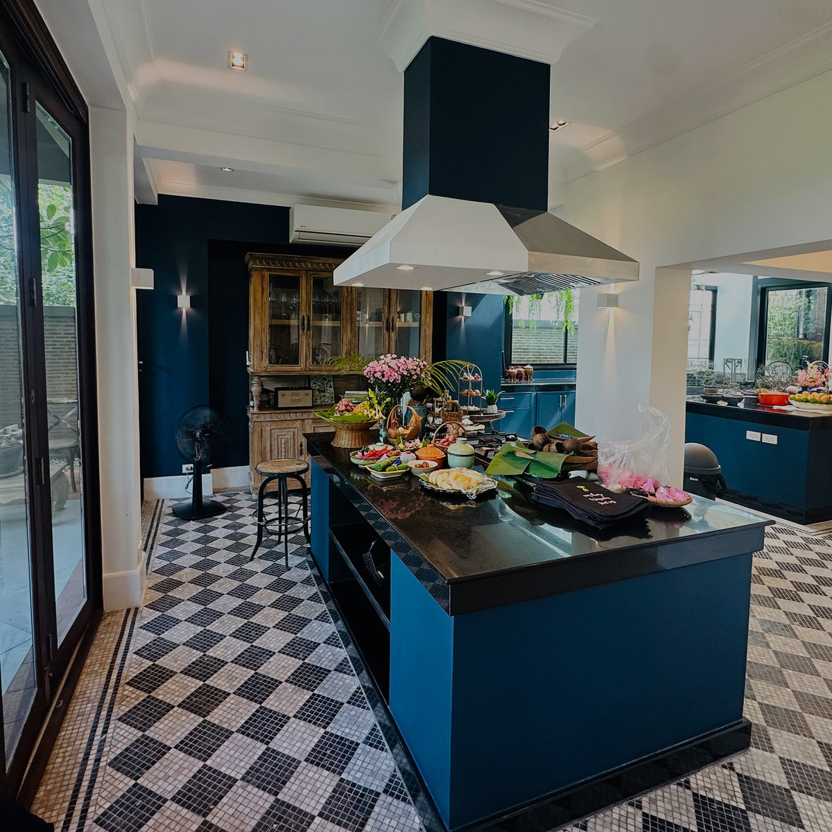 Sasi's kitchen interior with a prominent blue island and matching cabinetry, black countertops, a large white range hood, checkered black-and-white tile flooring, and decorative flowers and food on the island.