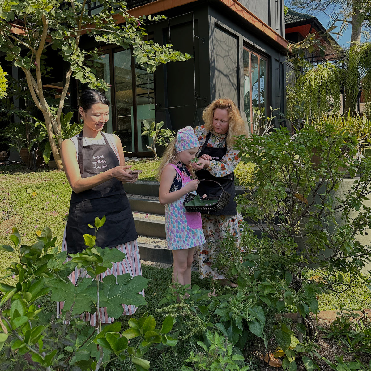Three women wearing black Sasi's Lovemade Kitchen aprons stand outdoors in a garden setting in front of a classic house, collecting herbs and vegetables from Sasi's herb garden.