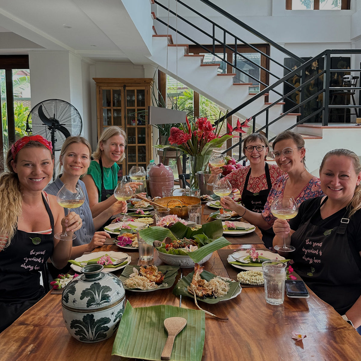 Seven women sitting around a dining table in a bright modern home, holding wine glasses and wearing matching black aprons with white text that reads 'The secret ingredient is always love.' The table is set with food, including tropical dishes, plates, and a decorative ceramic pot.