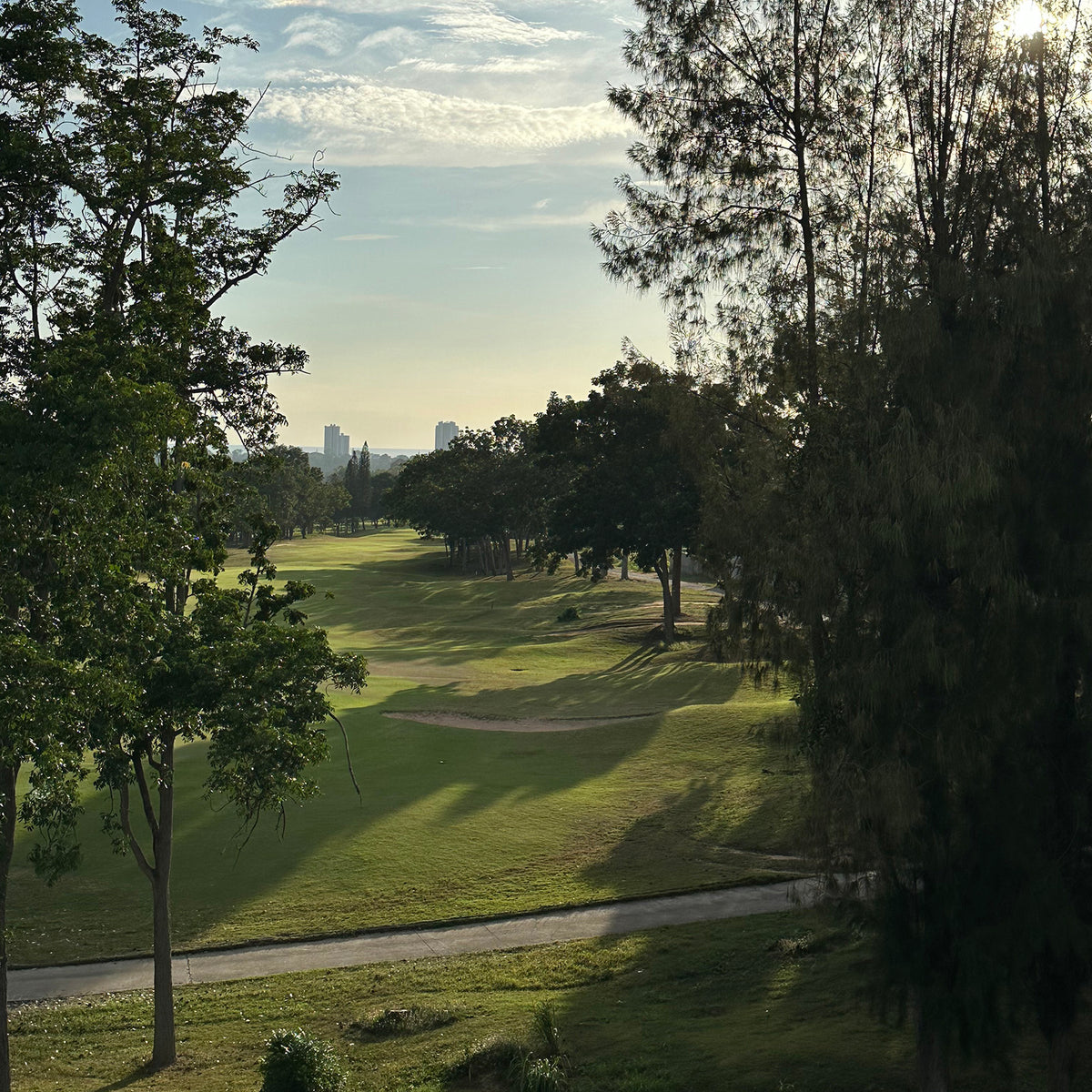 A scenic view of Phoenix Golf Course Pattaya from Sasi's Lovemade Kitchen's house with lush green grass, sand bunkers, and mature trees framing the shot under a bright sky with light clouds.