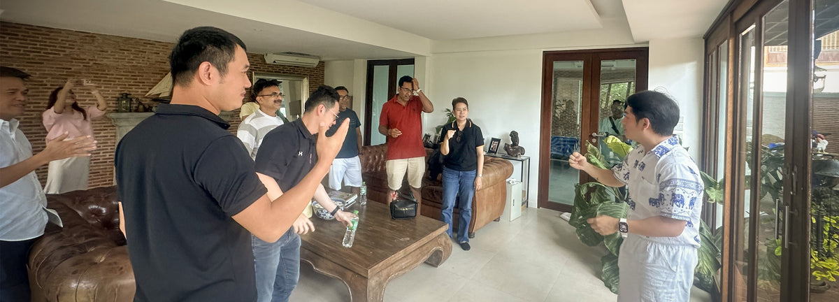 A group of men at a team-builing corporate event at Sasi's Lovemade Kitchen, standing and interacting inside a modern home living room with large glass doors, a brown leather sofa, and a wooden coffee table.