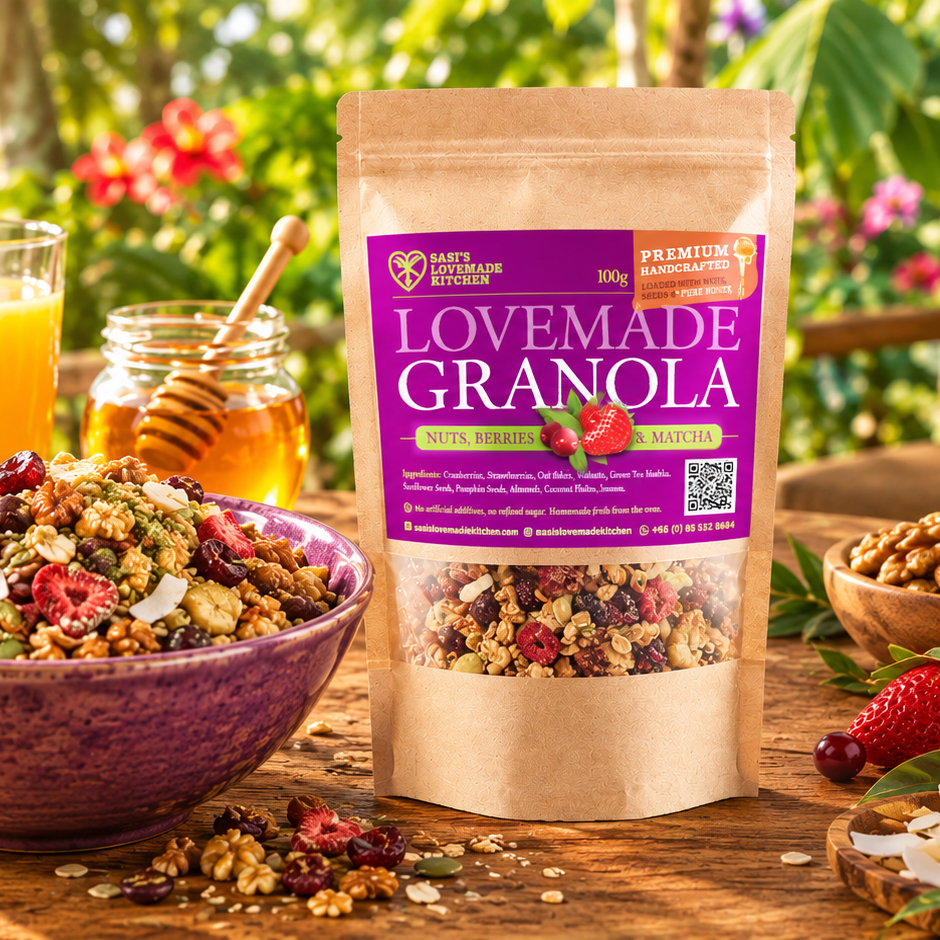 A 100 g bag of Granola by Sasi's Lovemade Kitchen stands on a wooden table outdoors, surrounded by a bowl of granola with strawberries and nuts, a jar of honey, and fresh strawberries. The product packaging is a kraft paper bag with a prominent purple label.